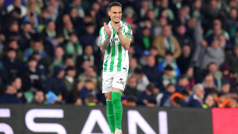 Brazilian footballer Antony, wearing a green and white Real Betis jersey, claps his hands while standing on the pitch during a La Liga match. The crowd in the background is filled with fans dressed in green and white.