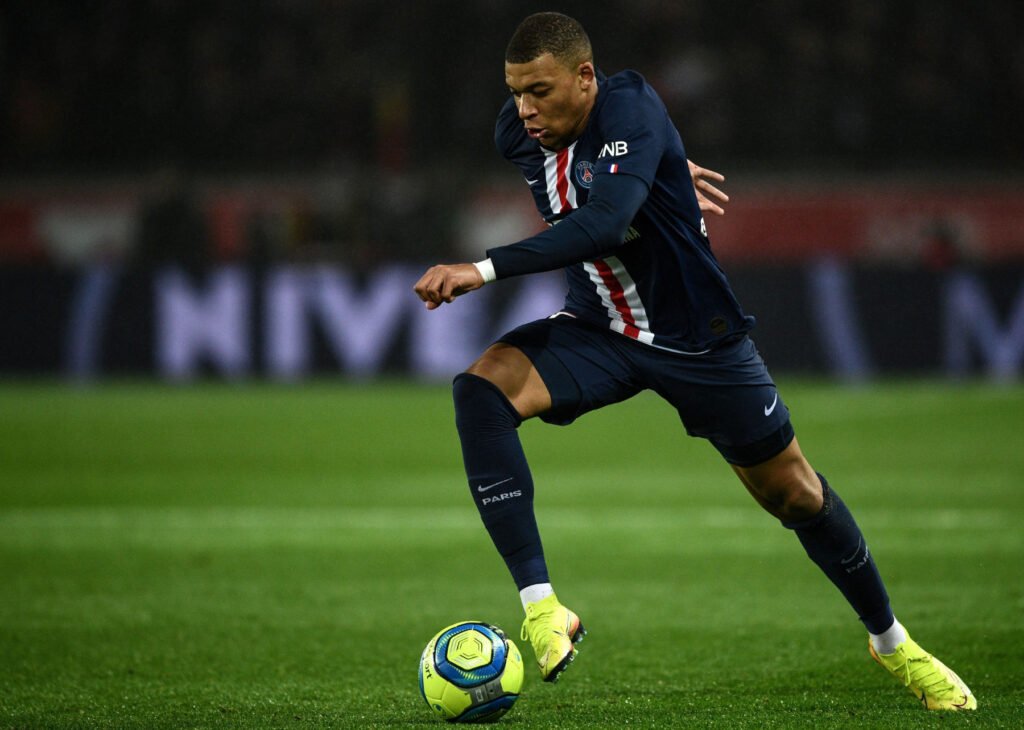 Kylian Mbappé celebrating with open arms during a football match, wearing the French national team jersey.