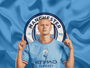 Erling Haaland's wearing a Manchester City jersey, standing in front of the club’s logo with both hands pointing upwards.