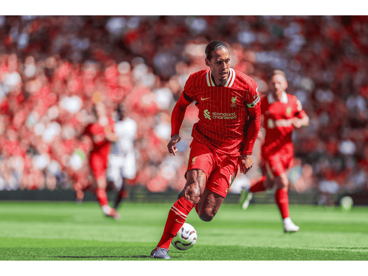 A Liverpool football player in red kit dribbles the ball on the field during a match, with a crowd in the background.