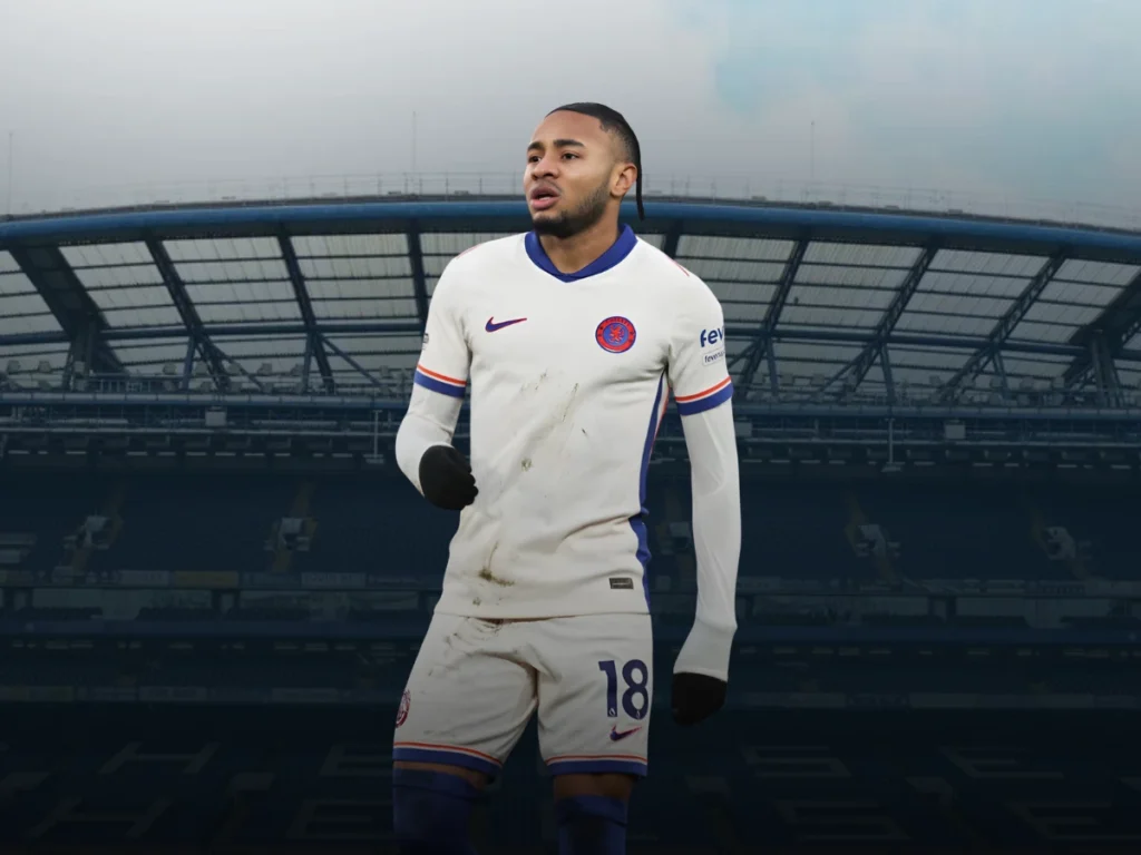 Christopher Nkunku wearing a white Chelsea FC away kit with the number 18 stands on the pitch at an empty stadium. The player has a focused expression, and his kit features subtle dirt marks, suggesting in-game action. 