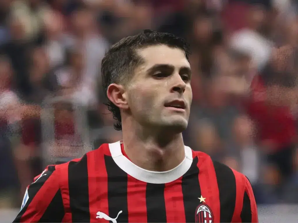 A football player wearing an AC Milan jersey with a focused expression, captured during a match with a blurred crowd in the background.