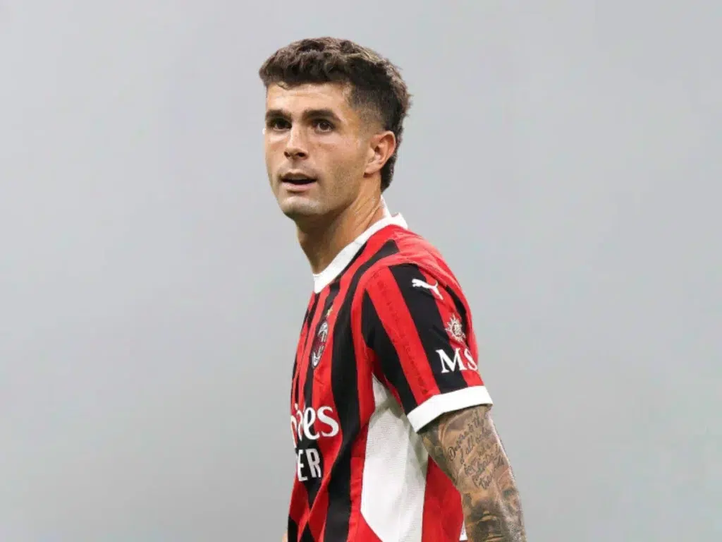 A football player wearing an AC Milan jersey with a focused expression, standing against a plain background, showcasing his tattoos and athletic physique.