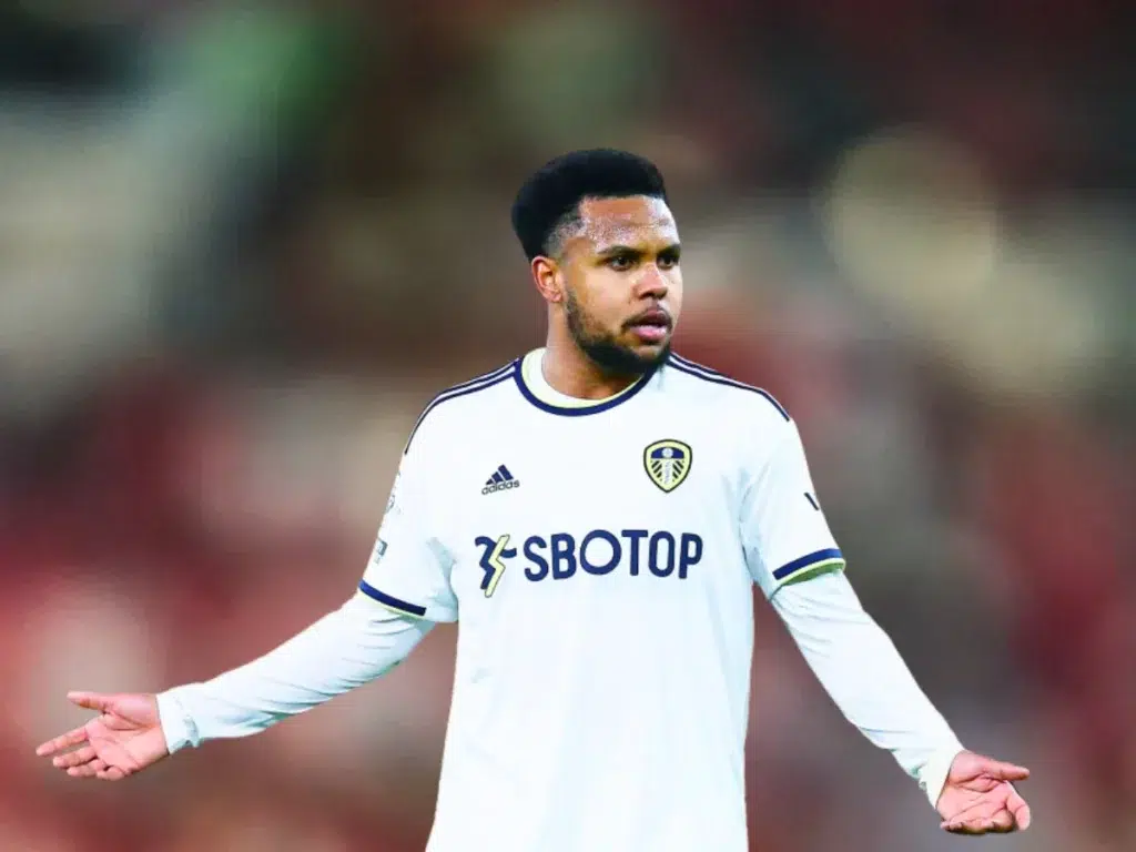 A soccer player wearing a white Leeds United jersey with the "SBOTOP" sponsor logo, standing on the field with his arms outstretched in a questioning gesture.
