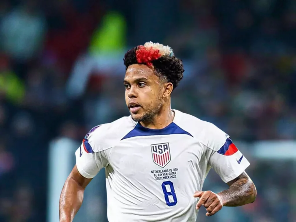 A soccer player wearing a USA national team jersey with the number 8, running on the field with a focused expression. His hair features a distinct red, white, and black color pattern.