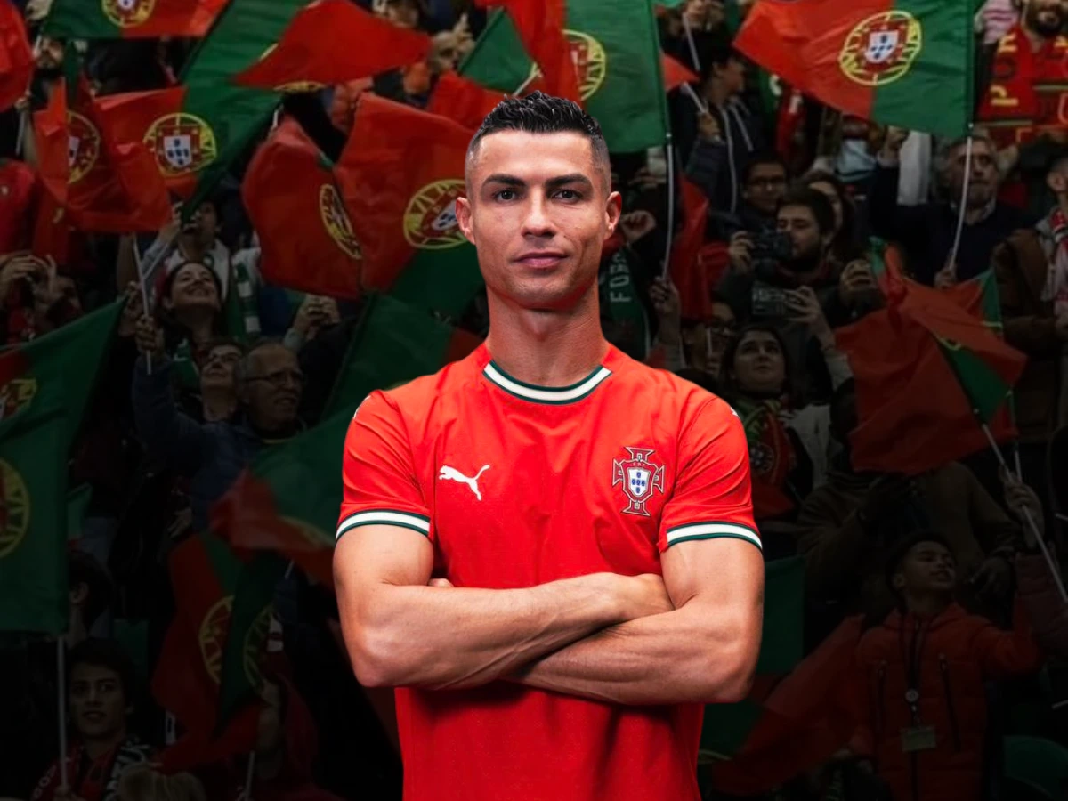 Cristiano Ronaldo stands confidently with his arms crossed, wearing Portugal's red national team jersey. Behind him, a crowd of passionate Portuguese fans waves green and red flags with the national emblem, creating an energetic and patriotic atmosphere.