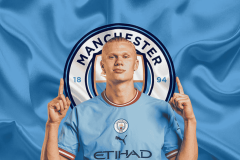 Erling Haaland's wearing a Manchester City jersey, standing in front of the club’s logo with both hands pointing upwards.