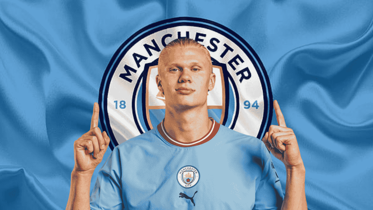 Erling Haaland's wearing a Manchester City jersey, standing in front of the club’s logo with both hands pointing upwards.