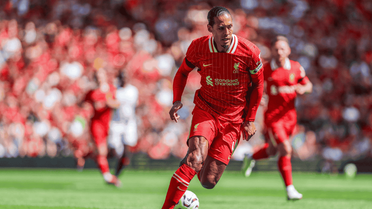 A Liverpool football player in red kit dribbles the ball on the field during a match, with a crowd in the background.