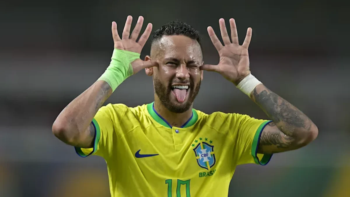 Neymar celebrating a goal while wearing Brazil's yellow jersey during an international football match, smiling and gesturing with his hands near his face.